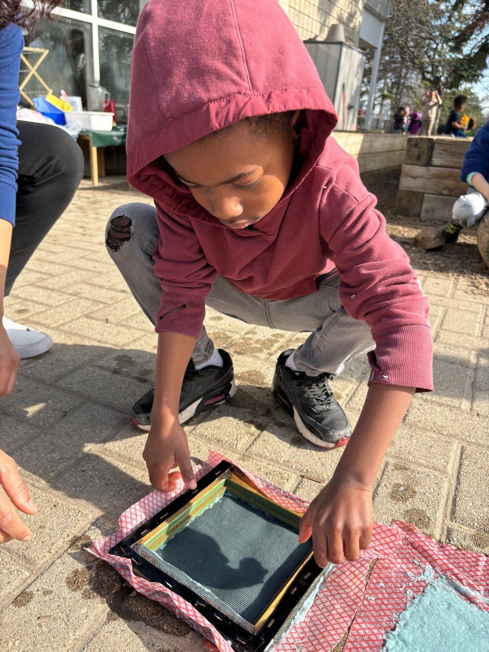 A young boy in a pink hoodie is kneeling down and looking at a piece of paper.