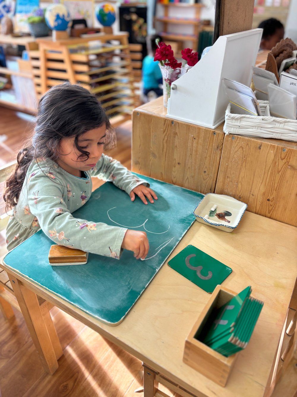 A little girl is sitting at a table playing with a chalkboard.