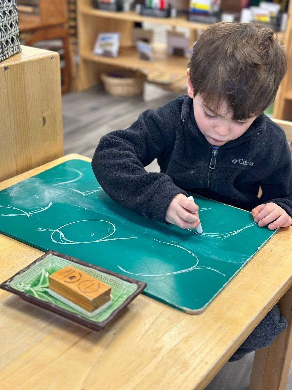 A young boy is sitting at a table writing on a chalkboard.