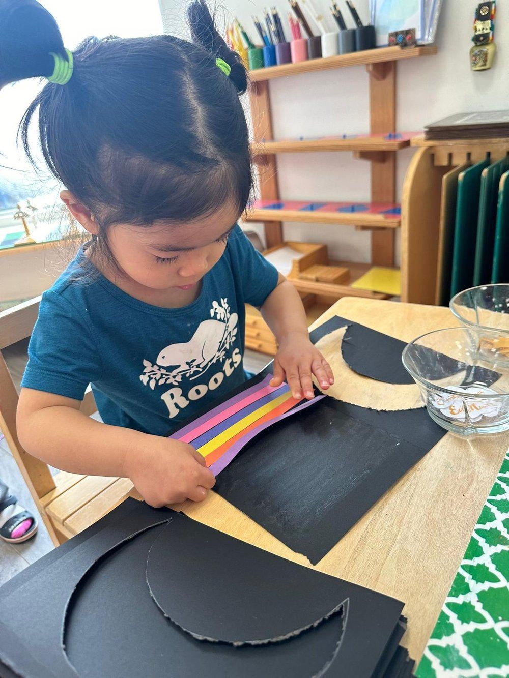 A little girl is sitting at a table making a rainbow out of paper.