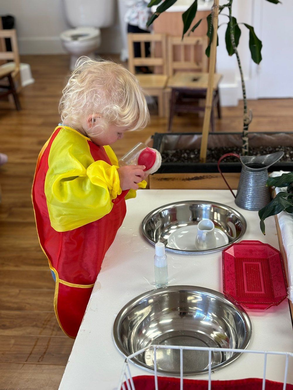 A little boy in a red apron is washing dishes in a kitchen sink.