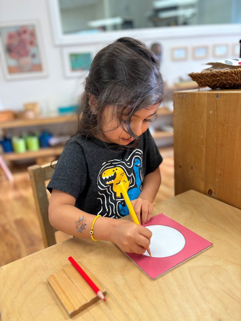 A little girl is sitting at a table drawing a circle with a pencil.