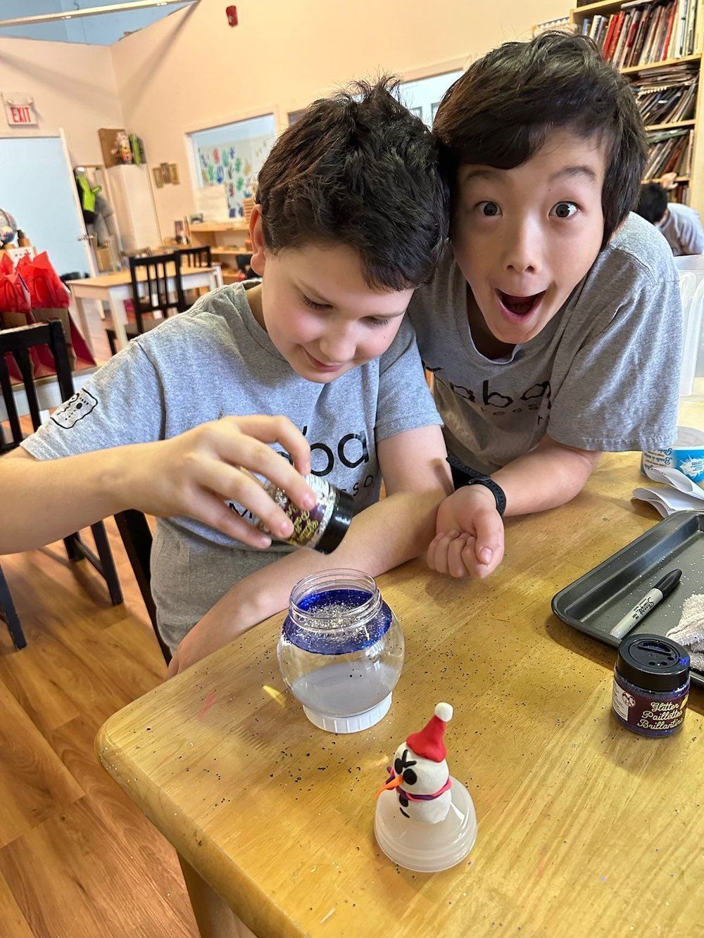 Two young boys are sitting at a table making a snow globe.