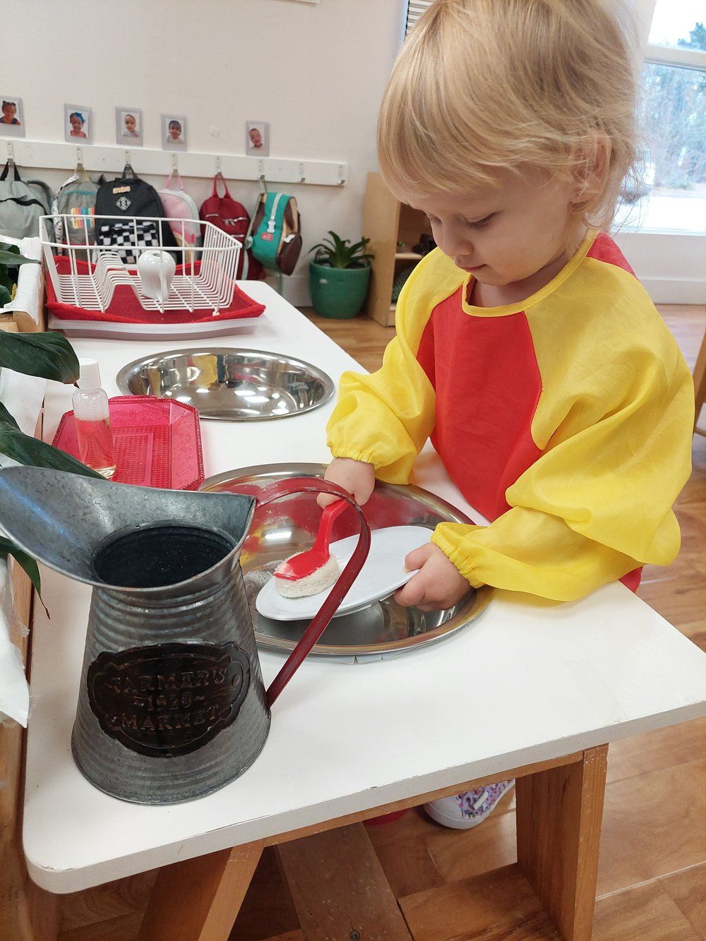 A little girl in a yellow apron is playing with a watering can on a table.