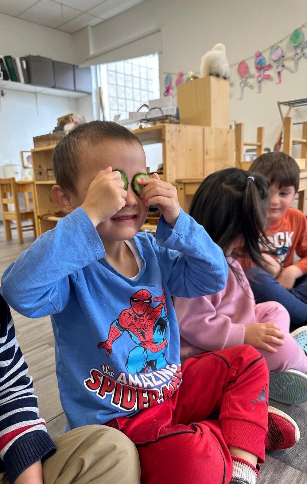 A young boy wearing a spiderman shirt is playing with cucumbers in a classroom.