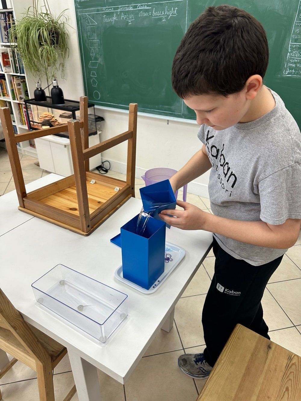 A young boy is standing at a table in a classroom holding a blue container.
