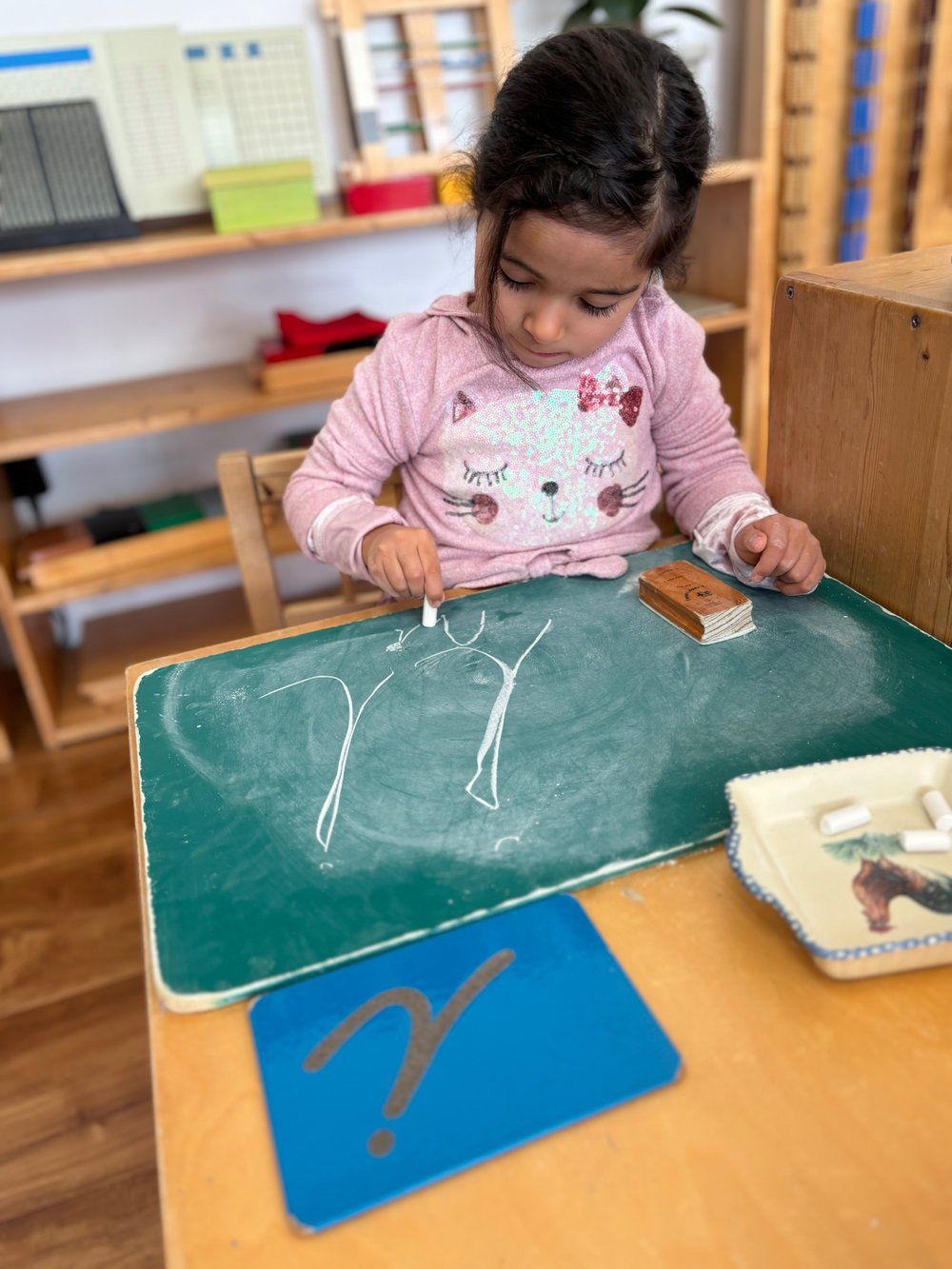 A little girl is sitting at a table writing on a chalkboard.