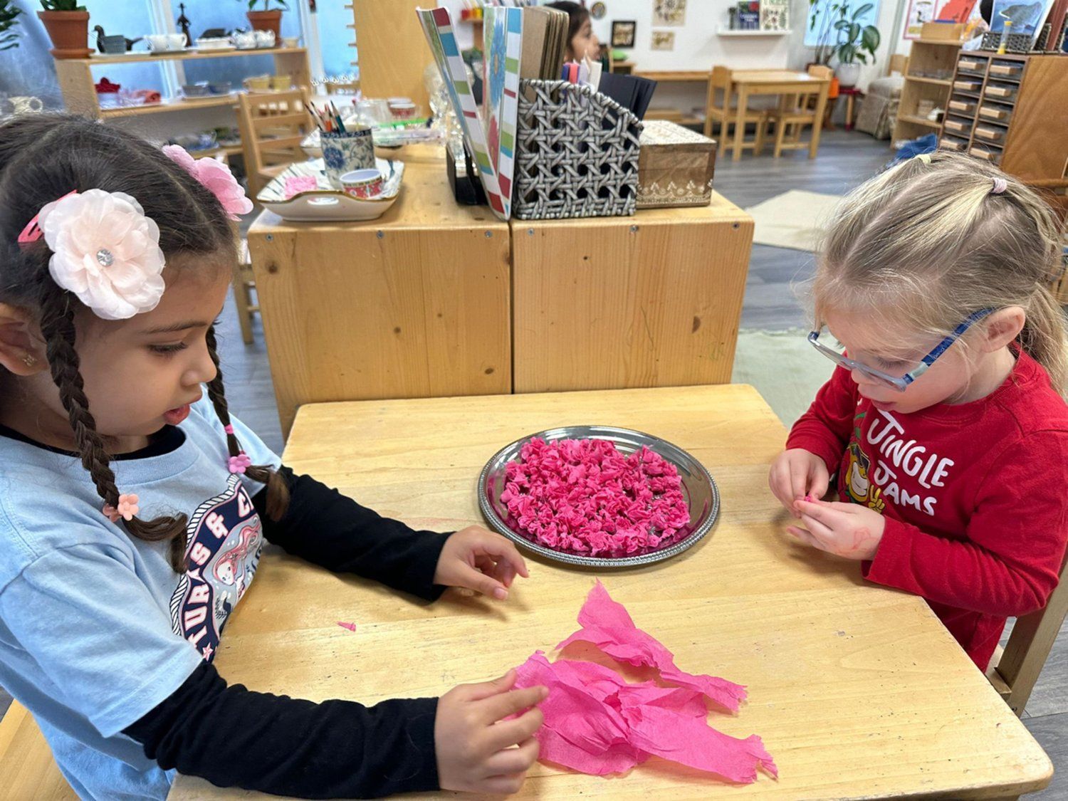 Two young girls are sitting at a table playing with pink tissue paper.