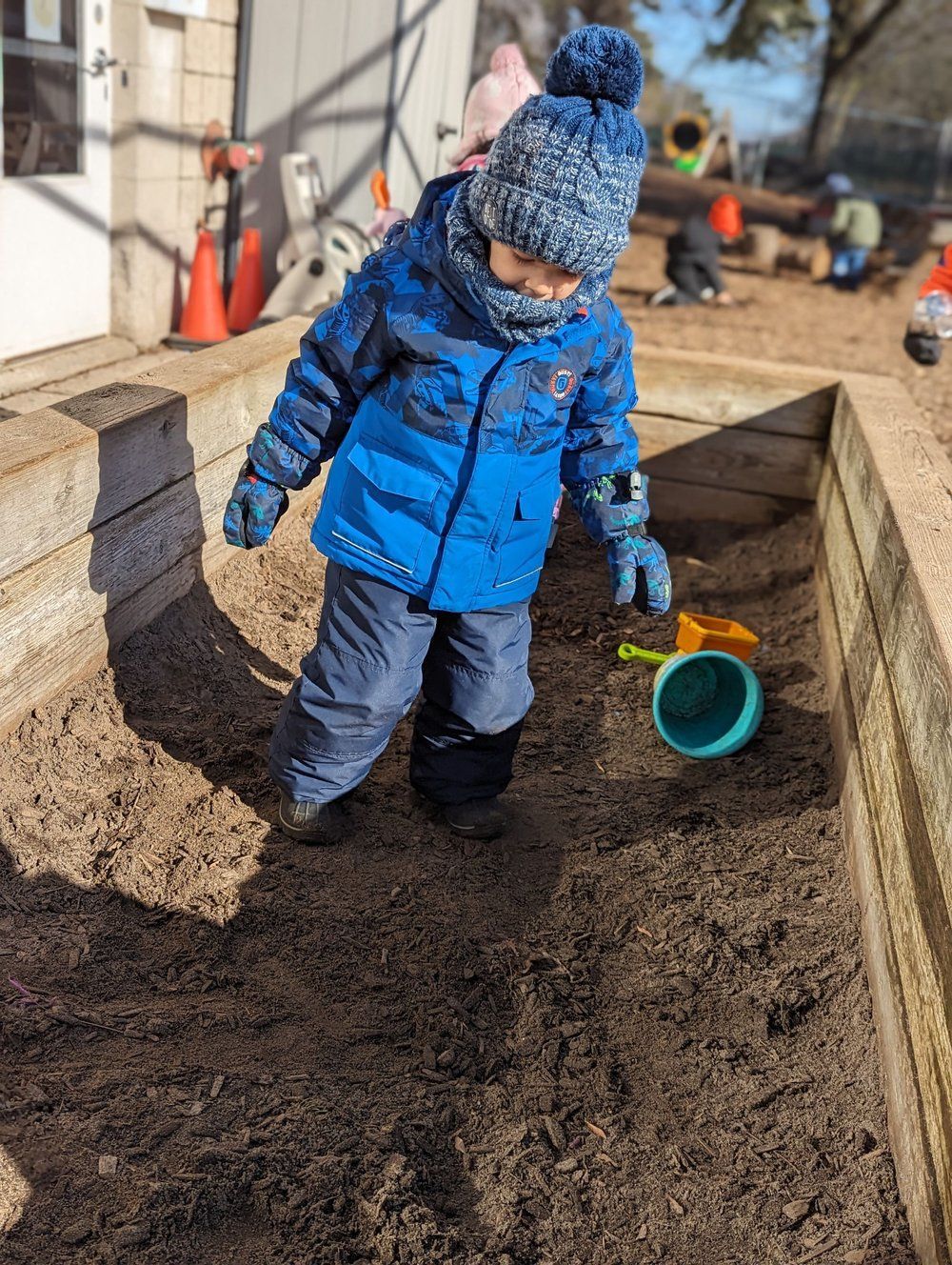 A little boy in a blue jacket and hat is playing in a sandbox.