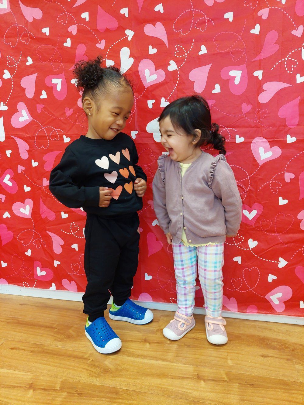 Two little girls are standing next to each other in front of a wall with hearts on it.