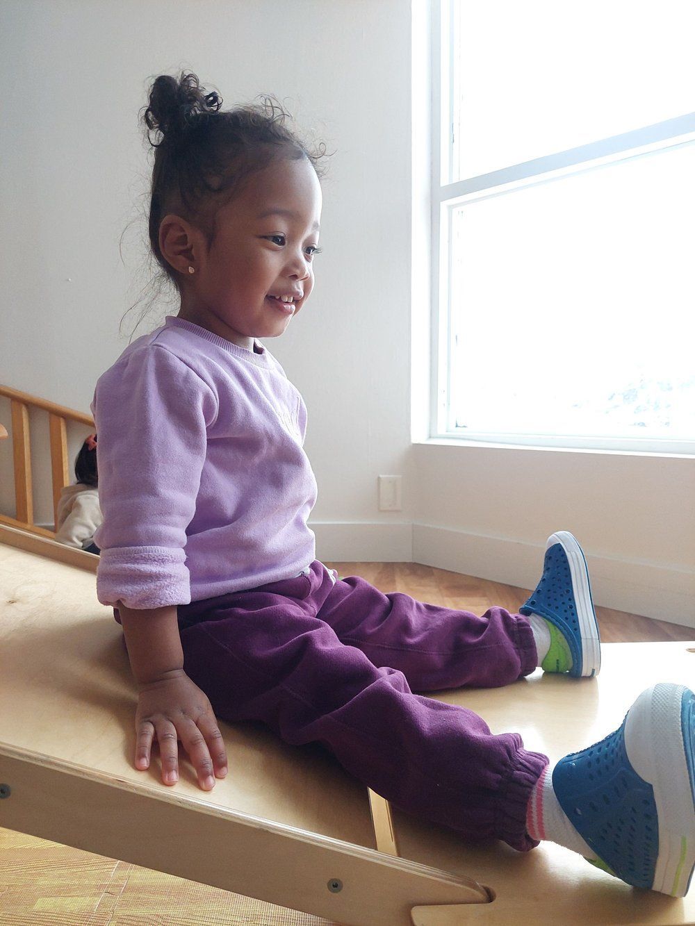 A little girl is sitting on a wooden slide and smiling.