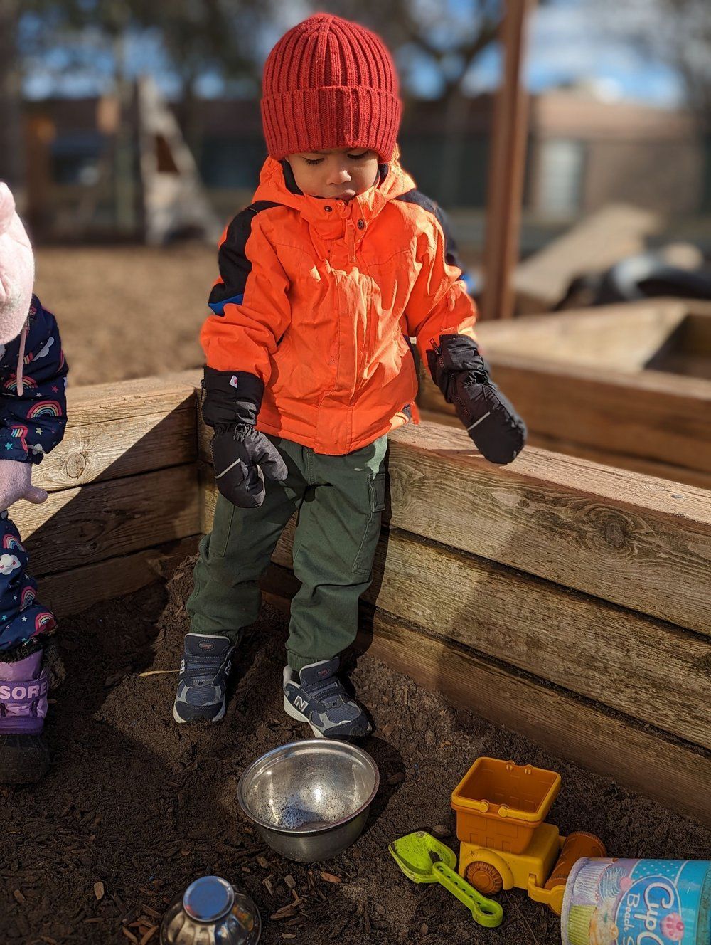 A little boy in an orange jacket is playing in a sandbox.