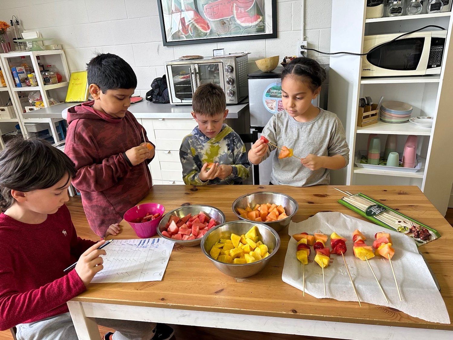 A group of children are sitting at a table making fruit skewers.