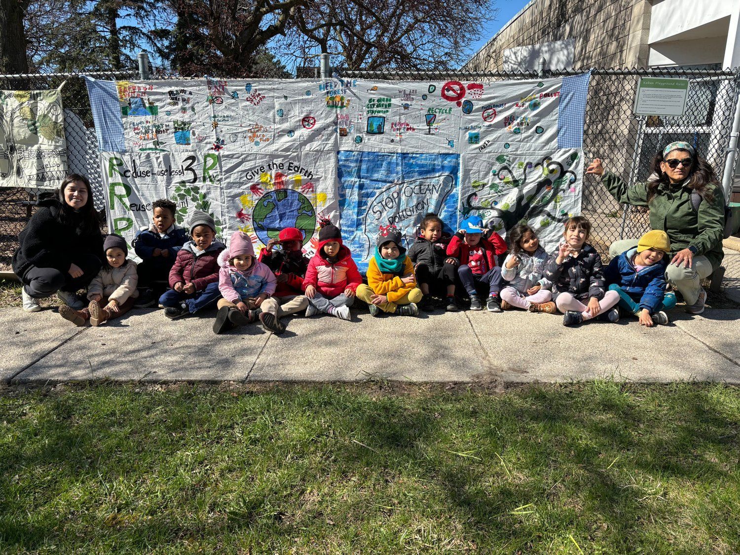 A group of children are sitting on the sidewalk in front of a fence.