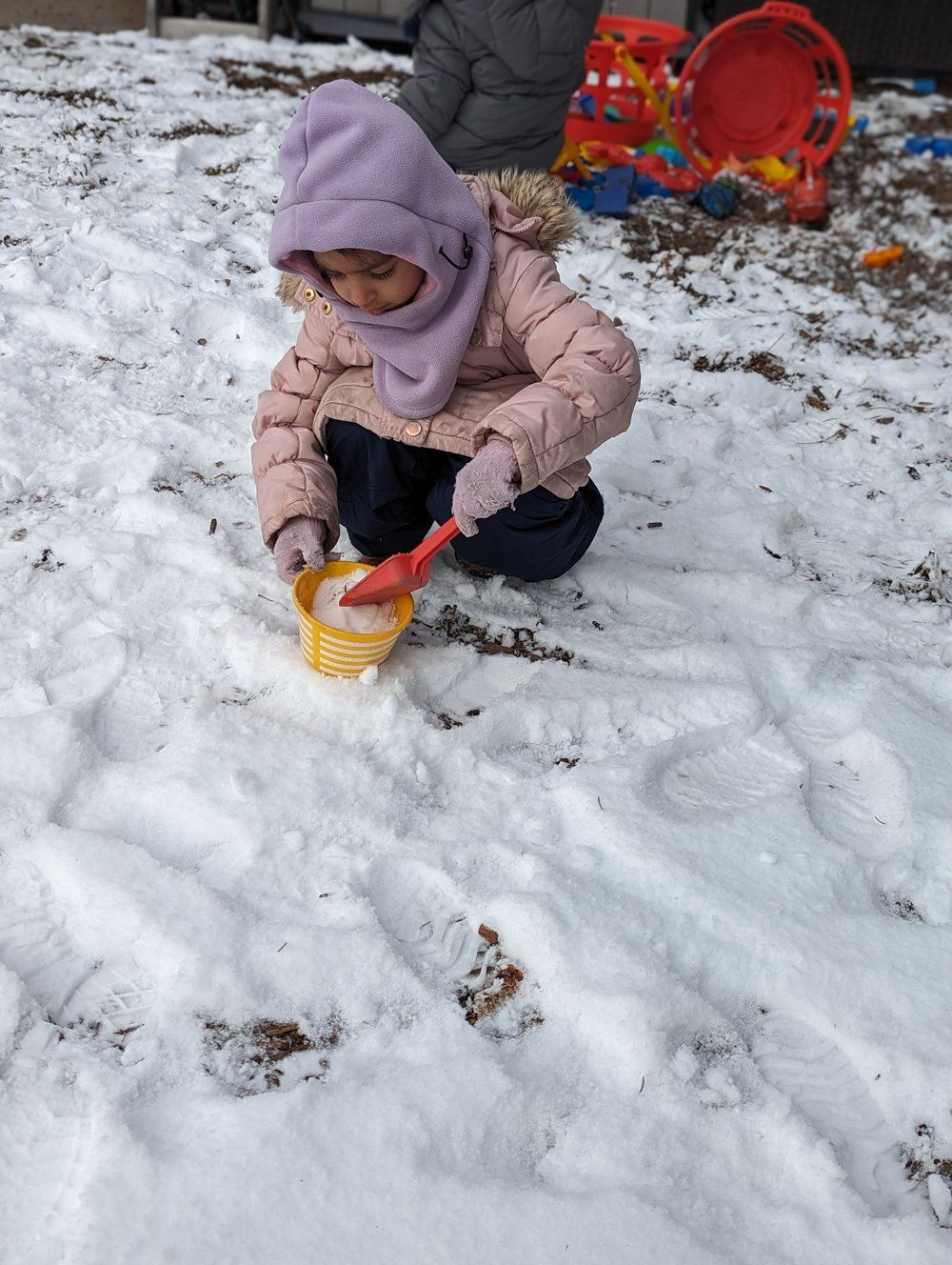 A little girl is playing in the snow with a yellow bucket