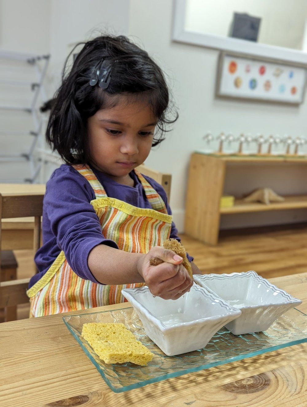 A little girl is sitting at a table playing with a sponge and bowls.
