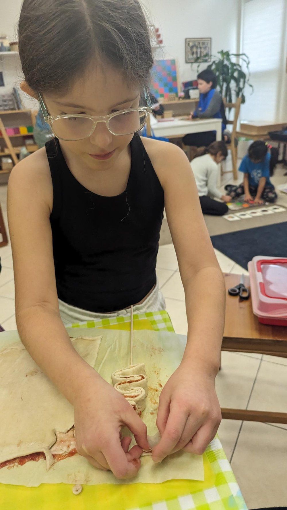 A young girl wearing glasses is making a sandwich in a classroom.