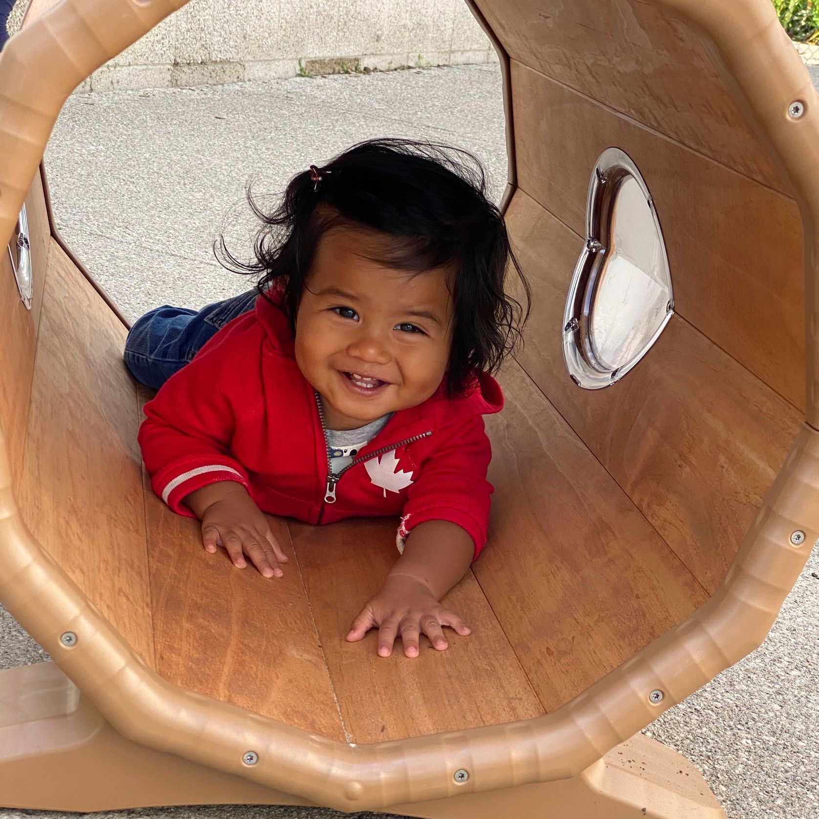 A baby in a red jacket is crawling through a wooden tunnel