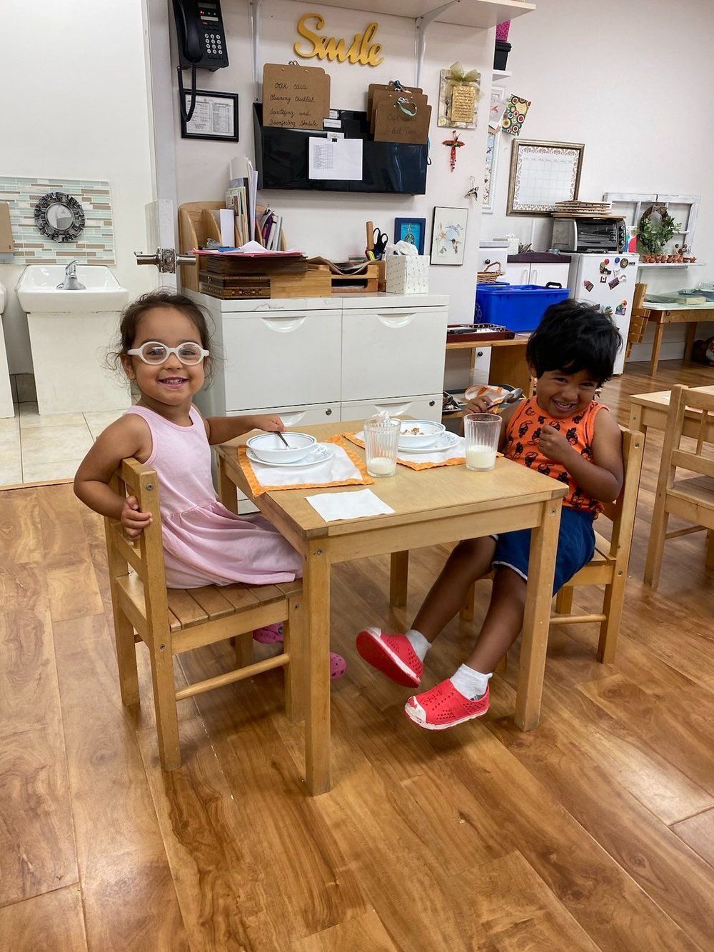 Two children are sitting at wooden tables in a classroom.