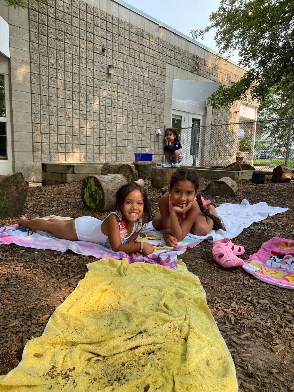 Two little girls are laying on a blanket in the dirt in front of a building.