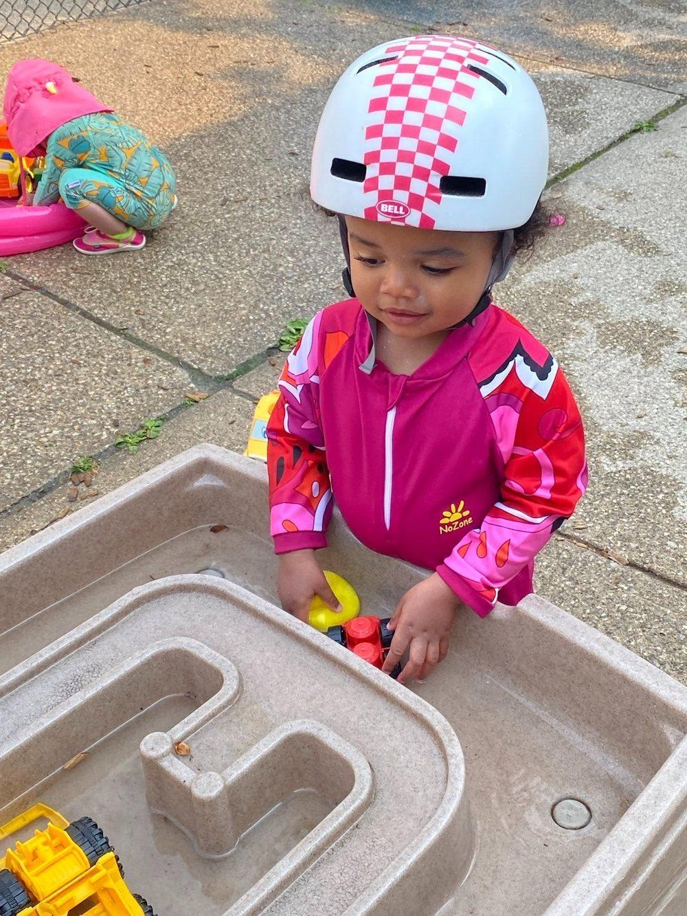 A little girl wearing a helmet is playing with toys in a water table.