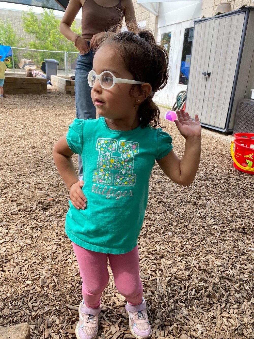 A little girl wearing glasses and a green shirt is standing in the dirt.