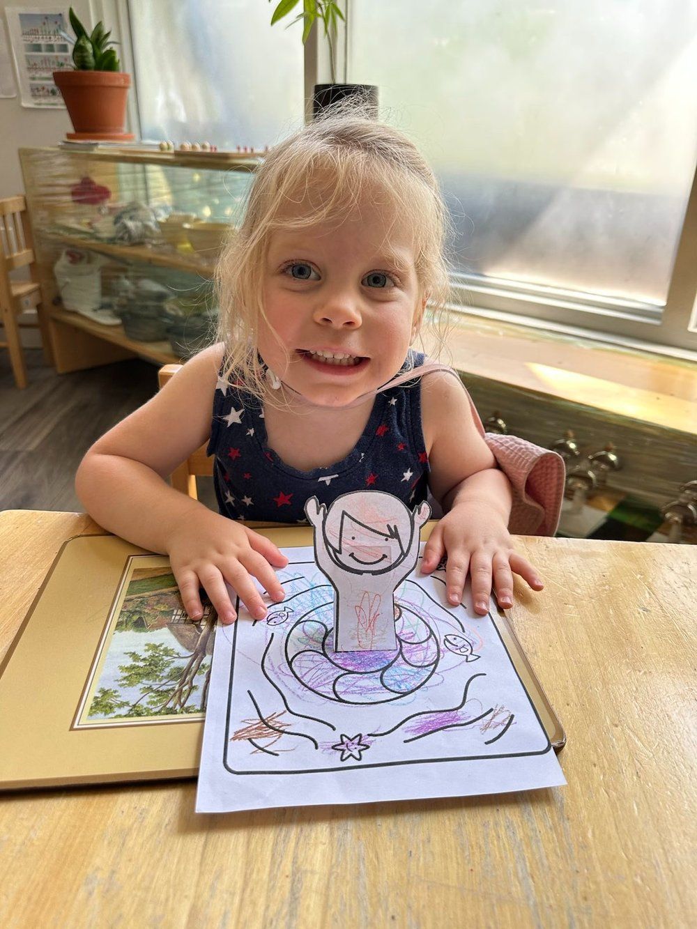 A little girl is sitting at a table with a drawing of a person in a life preserver.