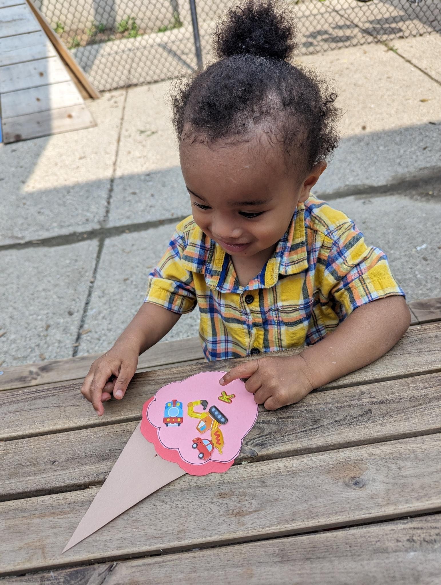 A little boy is sitting on a wooden table holding a paper ice cream cone.