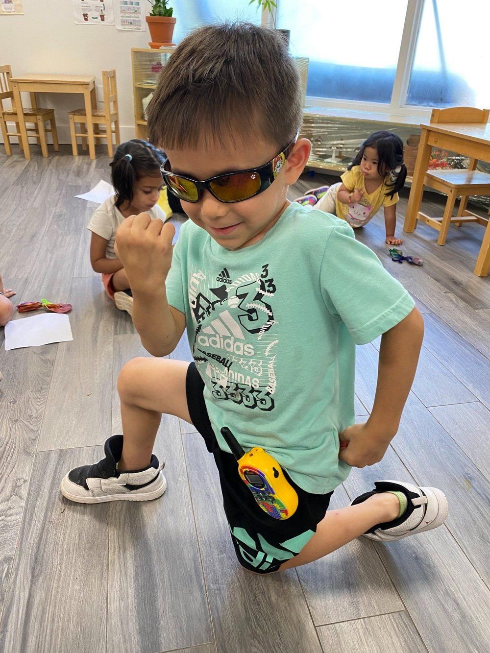A young boy wearing sunglasses is kneeling on the floor in a classroom.
