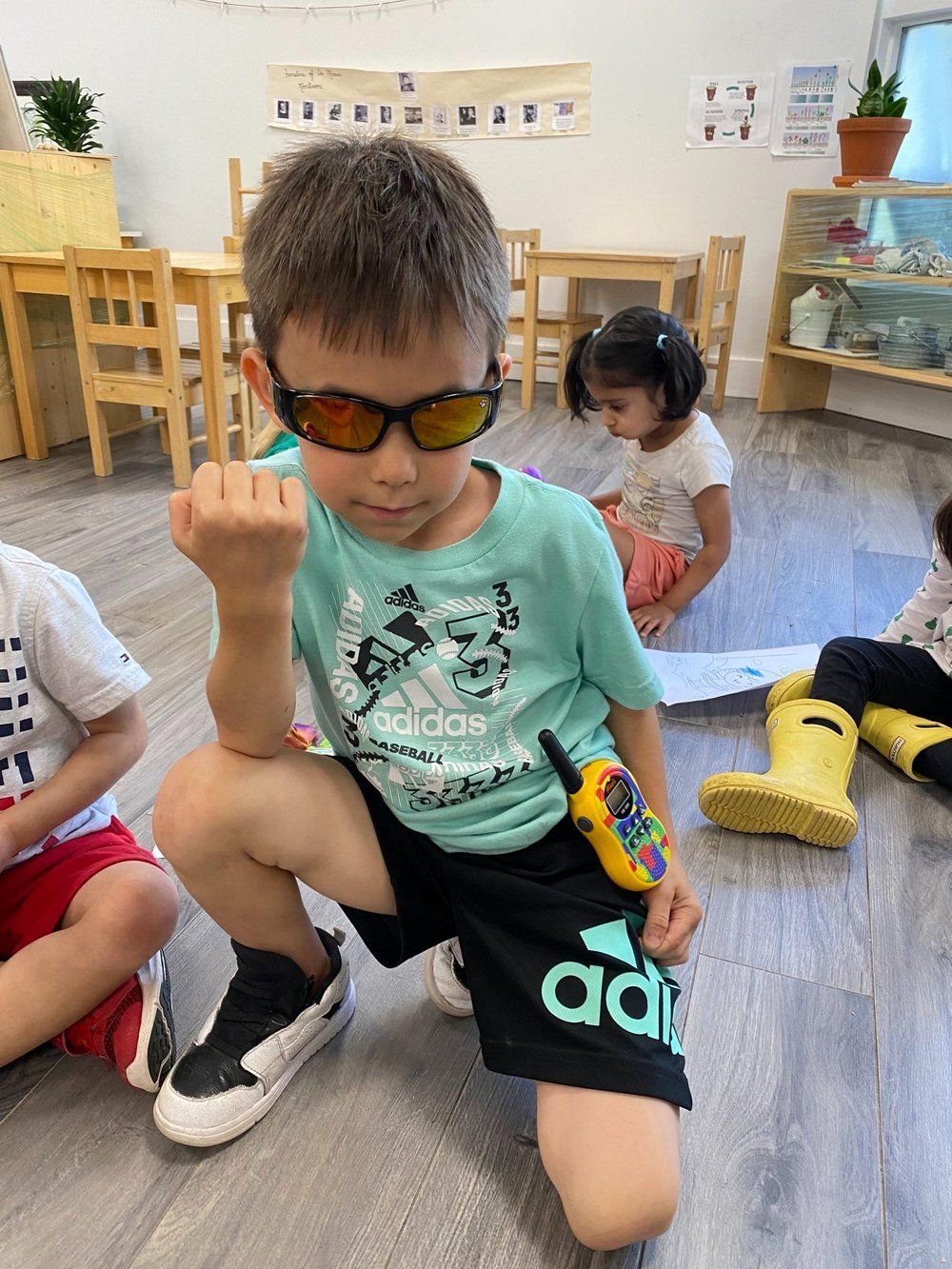 A young boy wearing sunglasses and a walkie talkie is kneeling on the floor.