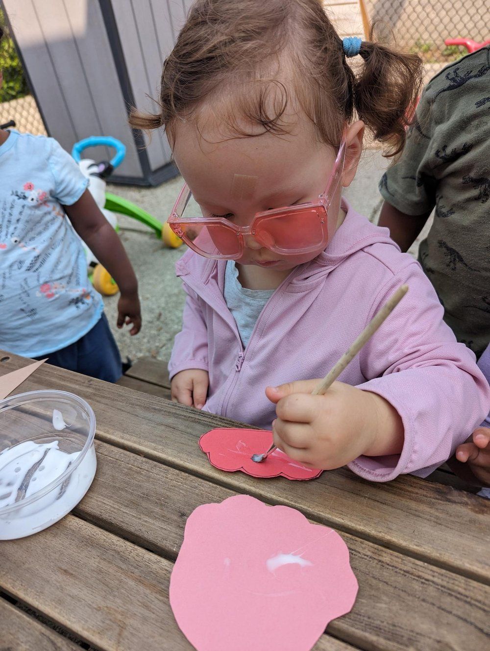 A little girl wearing sunglasses is painting a pink flower on a wooden table.