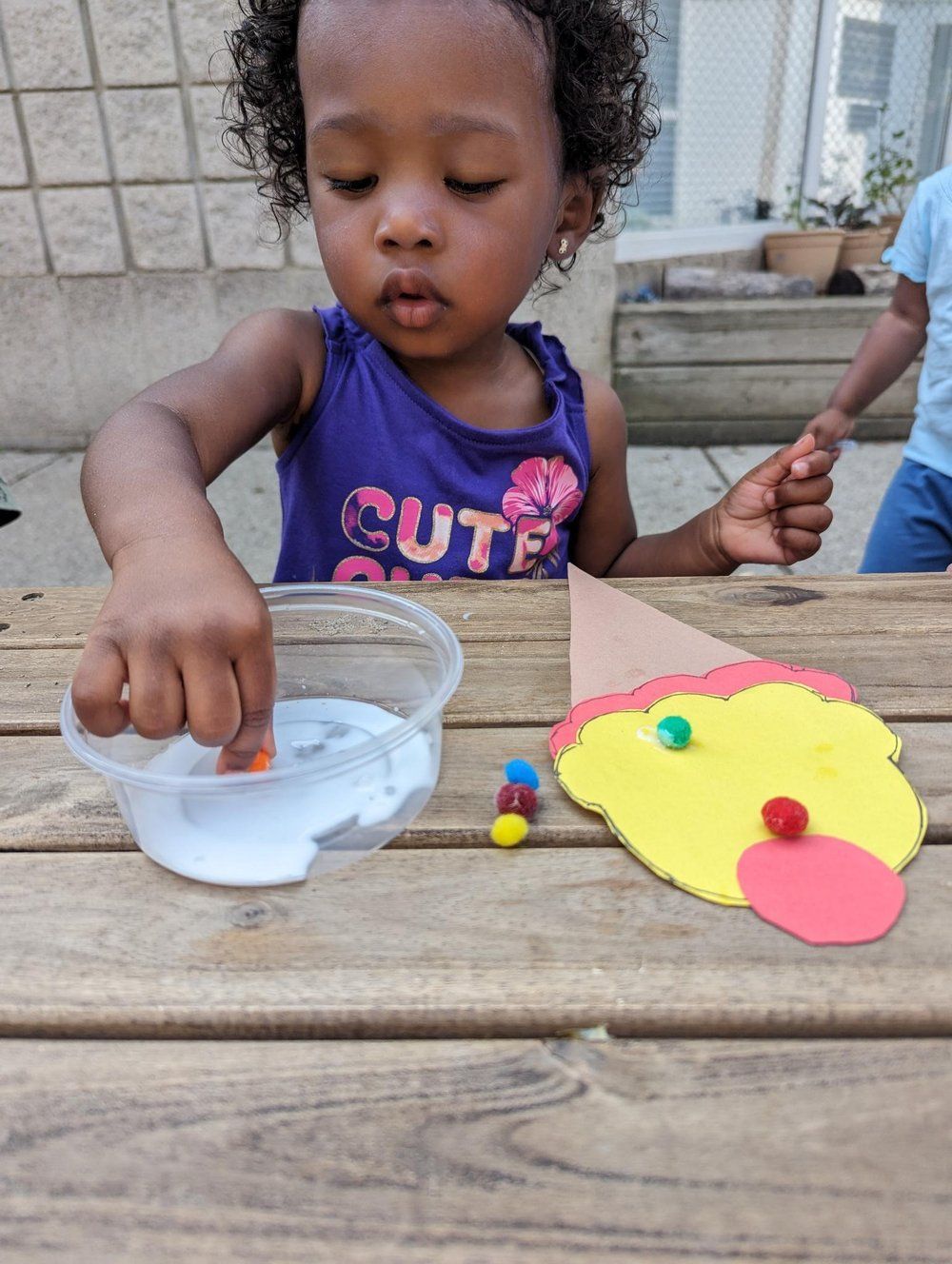 A little girl is sitting at a table making a clown face out of clay.