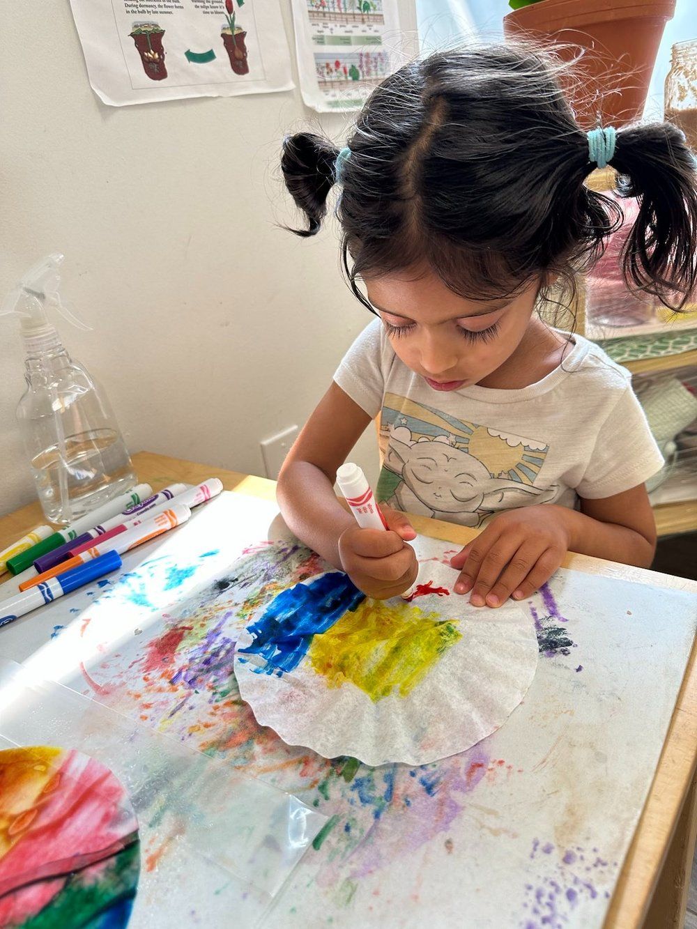 A little girl is sitting at a table painting on a paper plate.