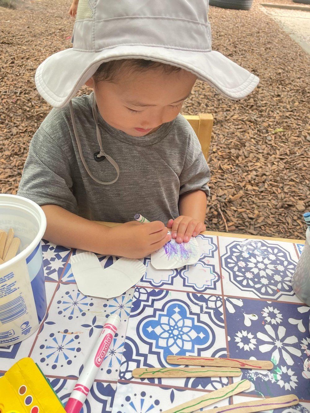 A little boy wearing a hat is sitting at a table drawing on a piece of paper.