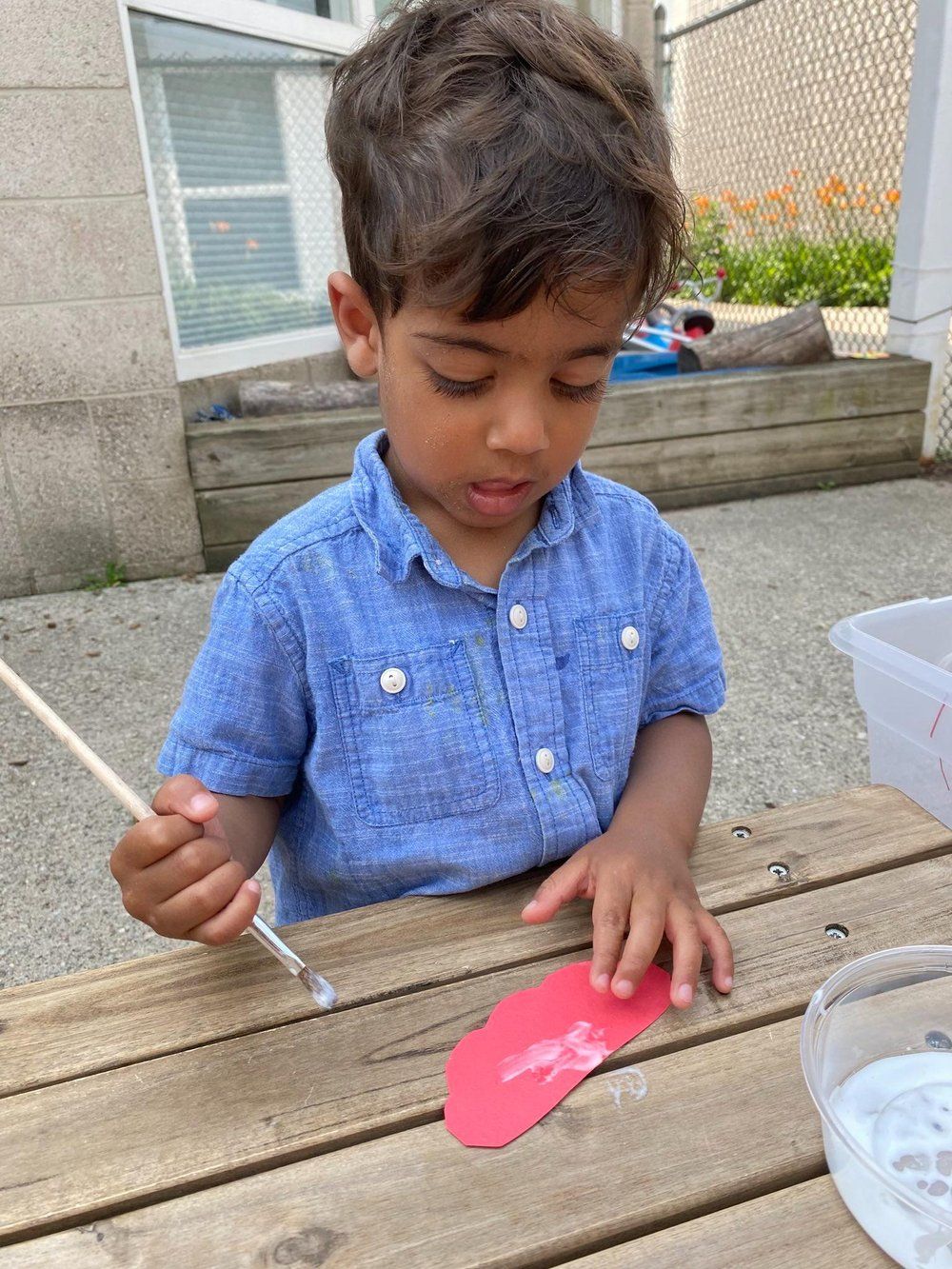 A young boy is sitting at a table painting a red object with a brush.