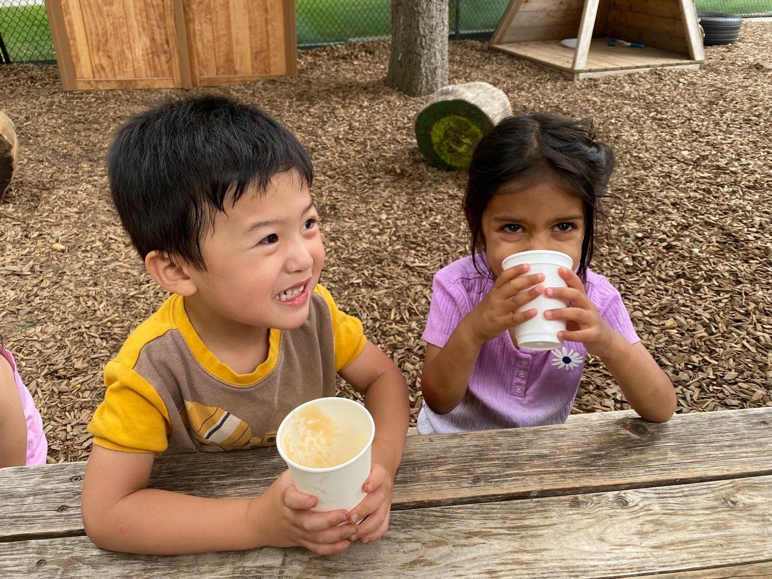 A boy and a girl are sitting at a picnic table drinking from cups.
