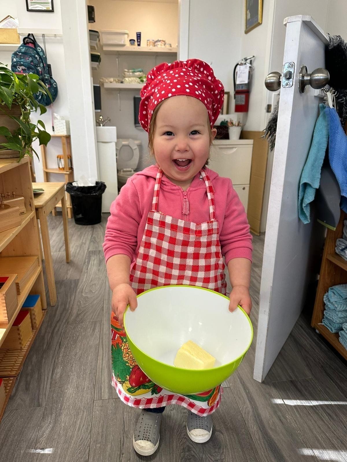 A little girl wearing a chef 's hat and apron is holding a bowl of butter.