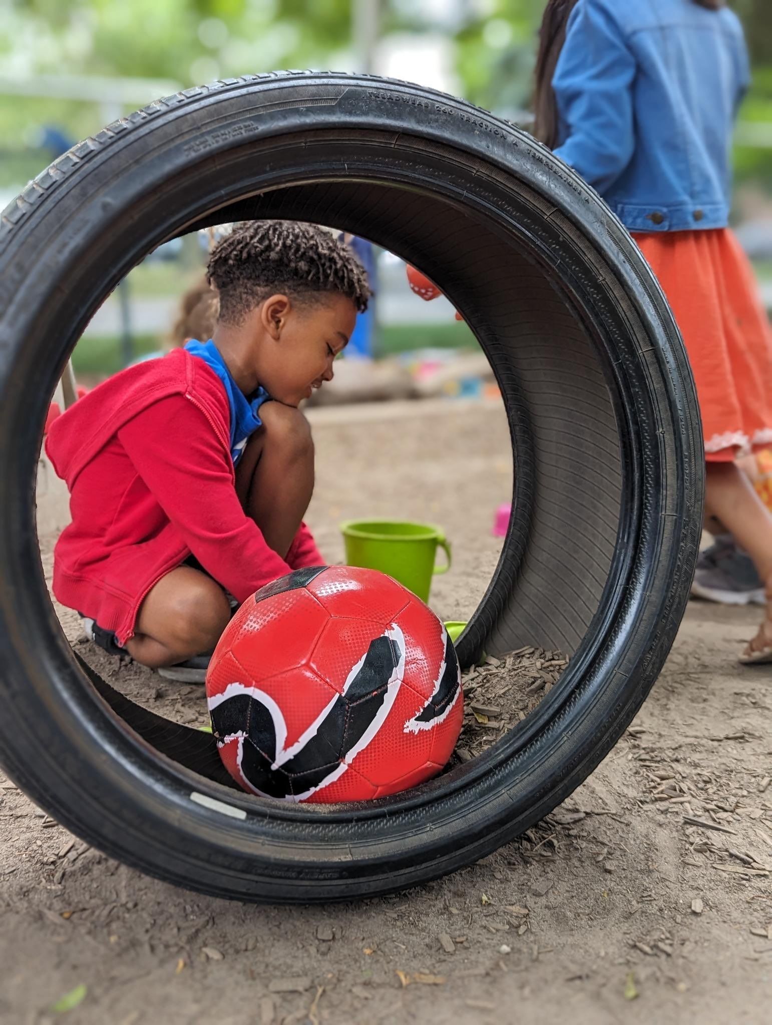 A young boy is playing with a red ball in a tire.