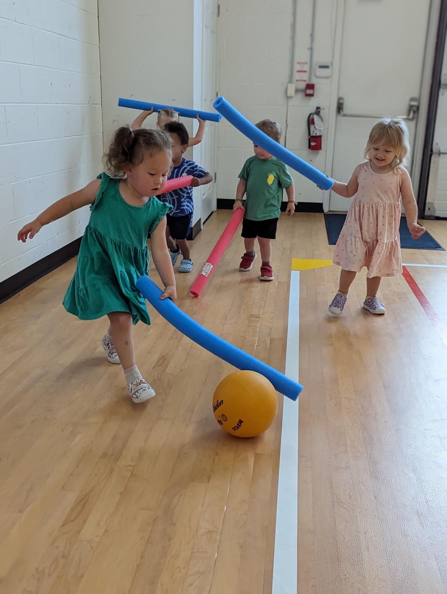 A group of young children are playing with pool noodles and a ball.