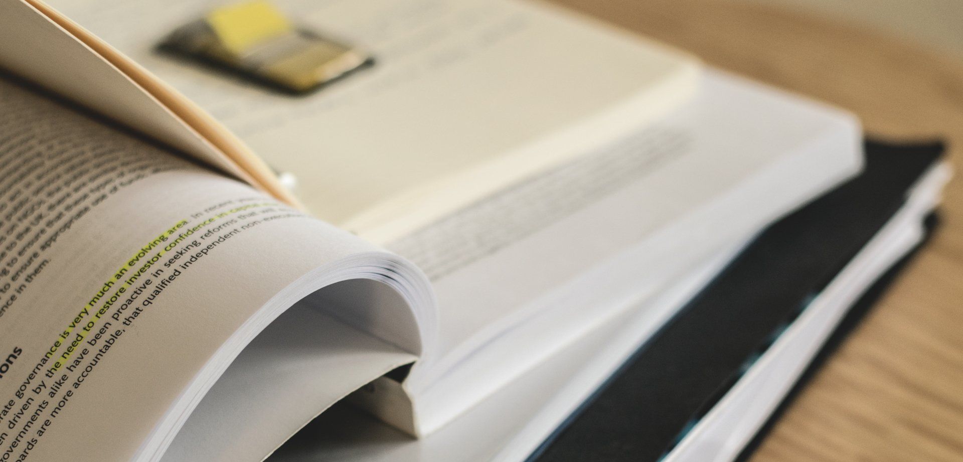 Open books with a highlighted passage and sticky note on top, stacked on a wooden surface.