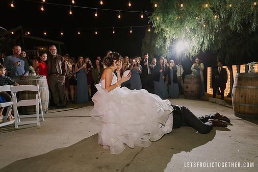 Bride sprawled on groom during reception dance, guests watching at night under lights.