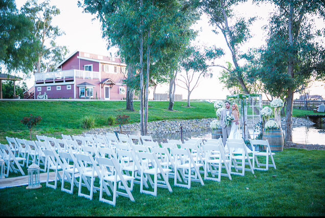 Wedding ceremony setup with white chairs on a grassy lawn; bride standing near floral arch, red building in background.