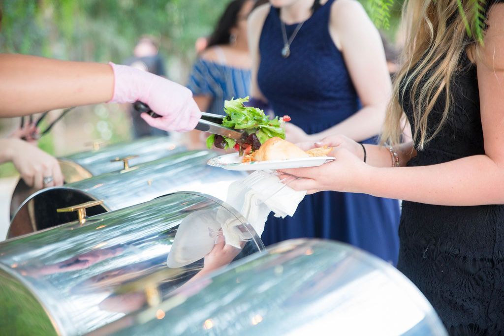 Person served food from a buffet with metal chafing dishes. Pink gloves handle tongs. Other people wait.