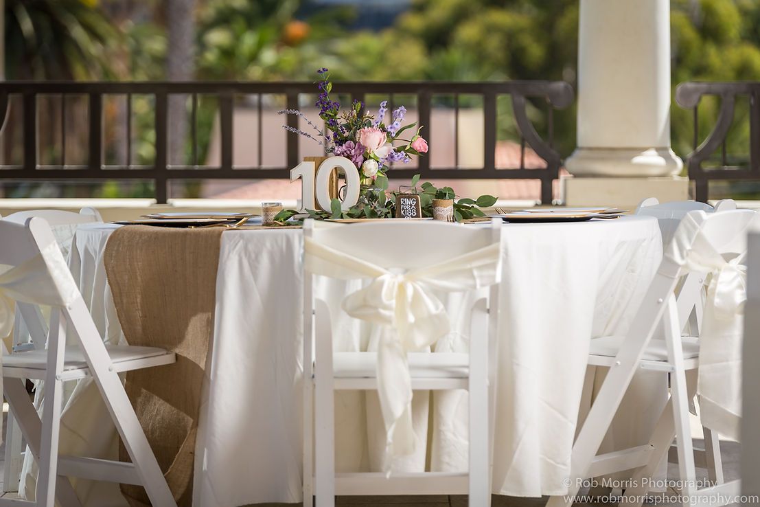 Wedding table with white chairs, draped in white linen and burlap runner, floral centerpiece.