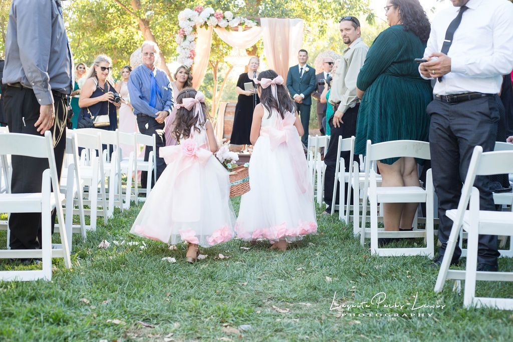 Two flower girls in pink dresses walk down an aisle at an outdoor wedding.
