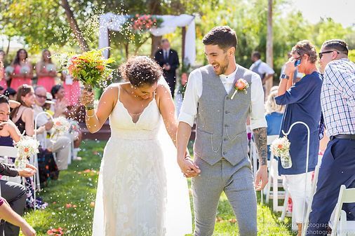 Newlyweds raise hands in celebration, cheered on by wedding guests, after ceremony by the sea.