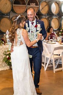 Wedding couple dancing, surrounded by guests. Money is pinned to groom's suit. Barrel backdrop, festive setting.