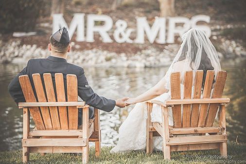 Couple in wedding attire holding hands, seated in wooden chairs by water with "MR & MRS" sign in background.