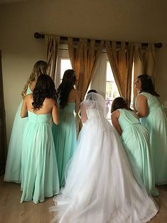 Bride in white gown with veil, surrounded by bridesmaids in mint green dresses, looking out a window.