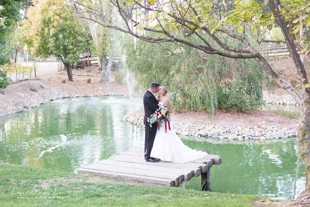 Couple kissing on a wooden dock by a pond, framed by a tree. Fountain and trees in the background.